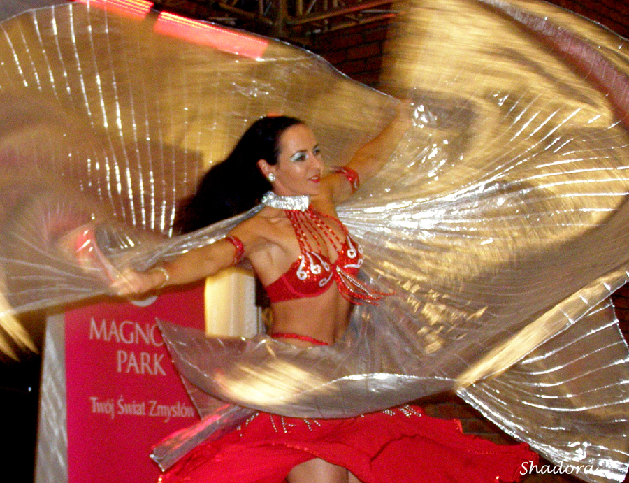 Tancerka w czerwonym, zdobionym stroju, prezentująca taniec orientalny z użyciem dużego, przezroczystego rekwizytu na tle sceny z napisem 'Magno Park'.